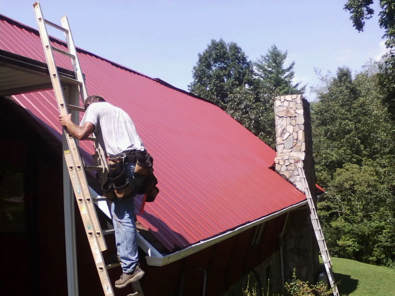 Roofer on a ladder beside a red metal roof and stone chimney — Soffit & Fascia Repair by Roofing Brothers in Stromsburg
